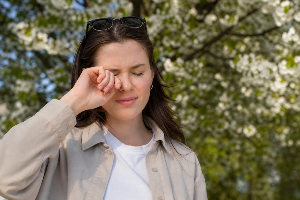 Person rubbing one eye outdoors near a tree, managing allergies.